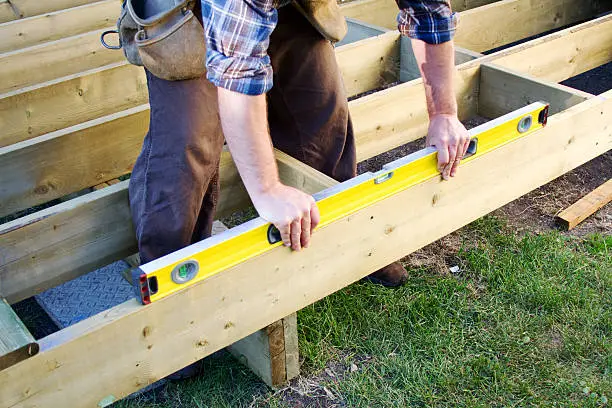 Skilled deck builder measuring framing layout on sloped backyard with precision tools
